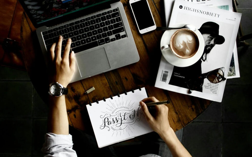 Hand sketching lettering in a notebook beside a laptop, coffee and a magazine