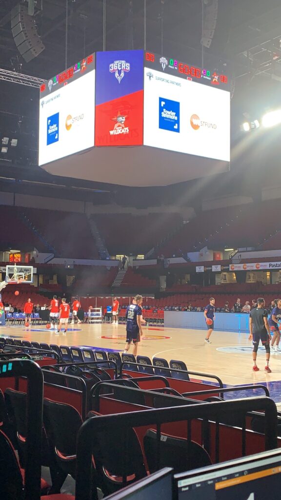 Centre-court screens showing sponsor content before an Adelaide 36ers game, with players warming up and a control laptop in the foreground