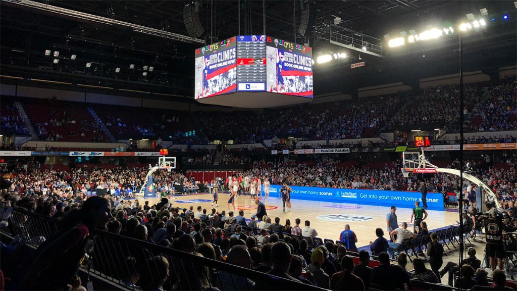 The four-sided centre-court screen above the court during an Adelaide 36ers game, showing a school holiday clinics promotion and a statistics table, with a sponsor message on the courtside LED boards