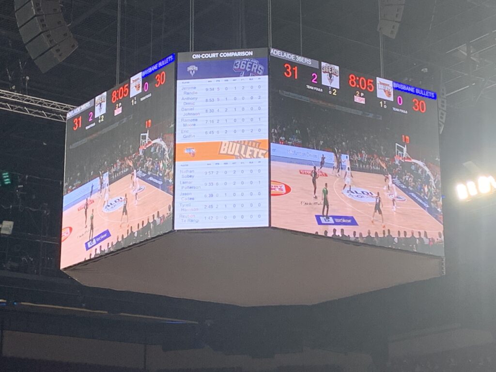 Centre-court screens at an Adelaide 36ers game showing an on-court statistics comparison and the score against the Brisbane Bullets