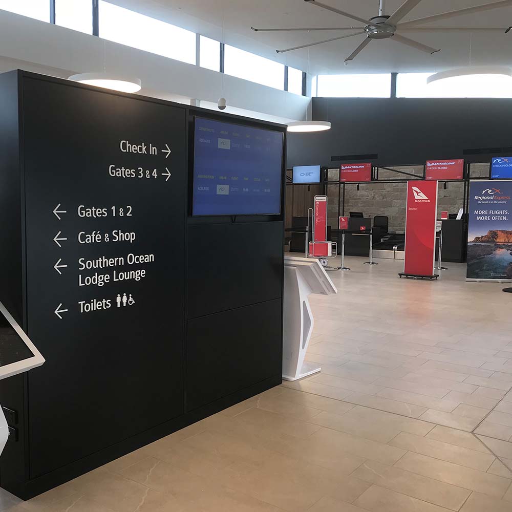 A flight information screen set into a wayfinding wall in the Kangaroo Island Airport terminal, with the check-in counters and their screens behind