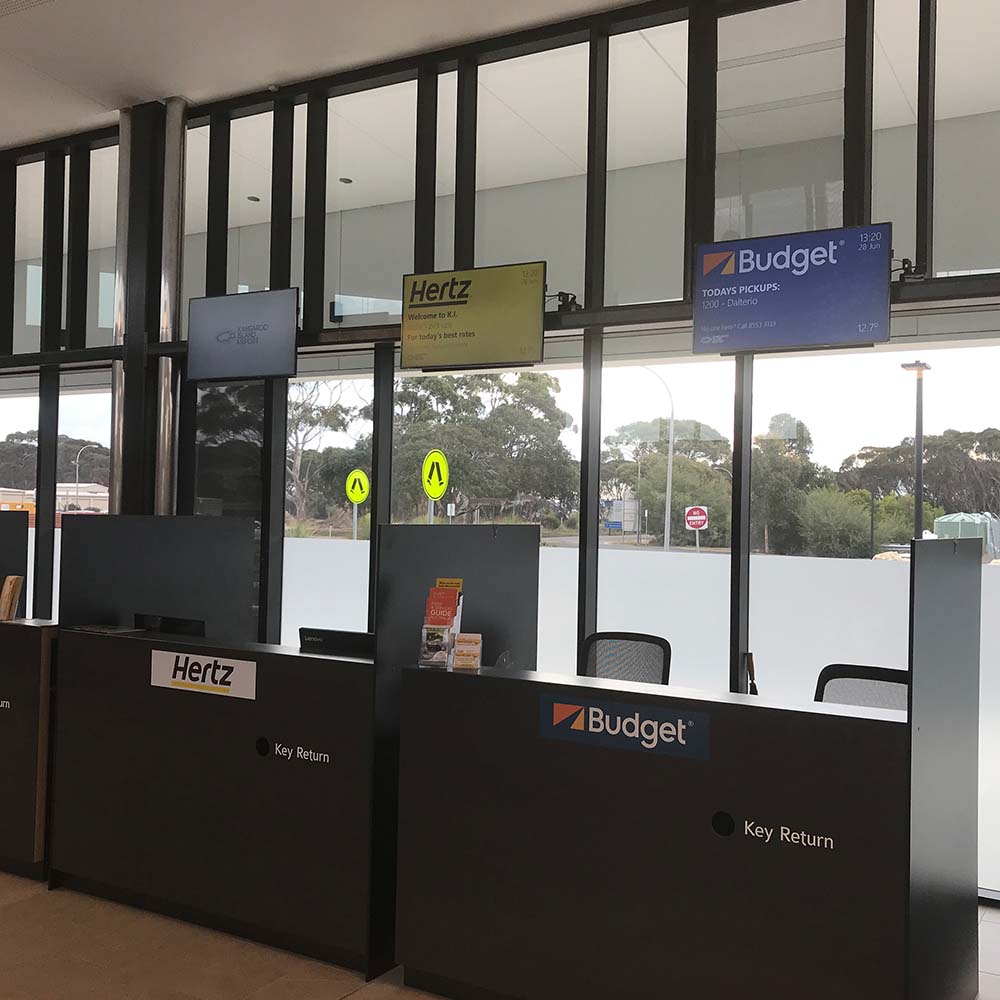 Screens above the Hertz and Budget hire-car desks at Kangaroo Island Airport, each in the company’s own colours, with the Budget screen listing today’s pickups