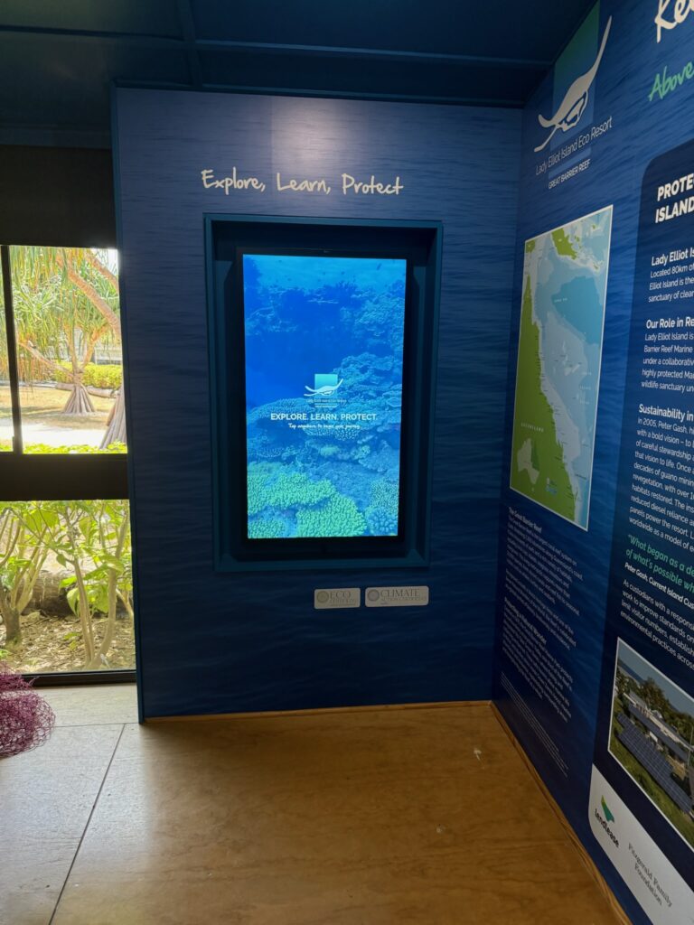 Portrait touchscreen set into a blue feature wall at Lady Elliot Island Eco Resort, showing reef imagery, the resort logo and “Explore. Learn. Protect. Tap anywhere to begin your journey”, beside a panel about the Great Barrier Reef